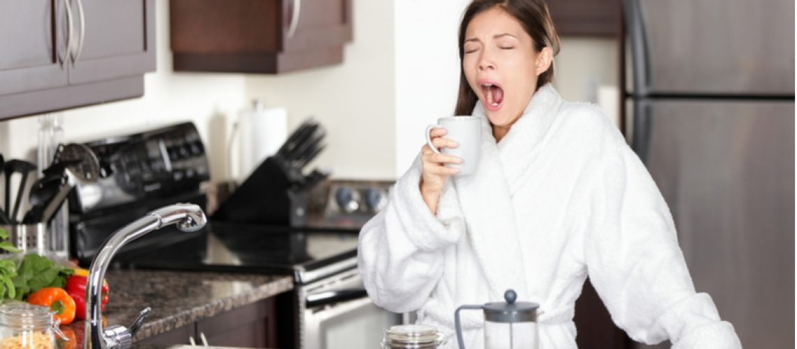 Woman in robe yawning in the kitchen while holding her coffee